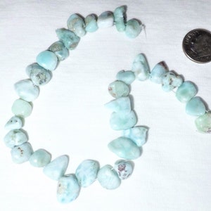 May include: Strands of light blue larimar gemstone beads. The beads are irregular in shape and size, with a mix of pale blue and white hues. A U.S. dime is next to the beads for size comparison. The beads are strung on a thin, white string.