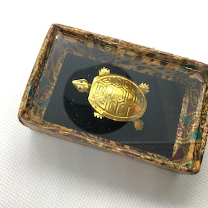 Puede incluir: Una pequeña figura de tortuga en tono dorado con un patrón de caparazón detallado, colocada dentro de una caja de presentación rectangular. La caja presenta un borde estampado en tonos marrón, dorado y verde, con una tapa transparente y una base de terciopelo negro.