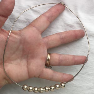 May include: A silver necklace featuring a thin, flexible chain with a series of round, metallic beads. The necklace is held up against a hand, showcasing its design and the way it might sit when worn. The clasp is visible.