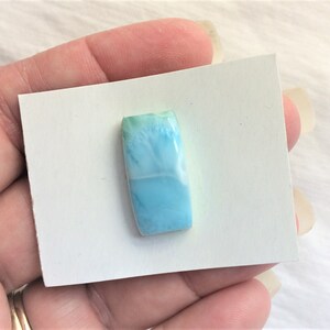 May include: A rectangular, polished gemstone with a light blue and white color gradient. The stone is held on a white card, showcasing its smooth surface and natural patterns. The gemstone is approximately 1 inch in length.