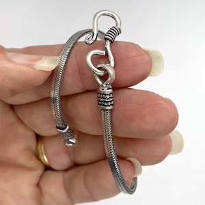 May include: A silver-toned snake chain bracelet featuring a textured herringbone pattern. The bracelet is secured with a prominent hook-and-eye clasp mechanism, which includes decorative ribbed metal detailing at the connection points.