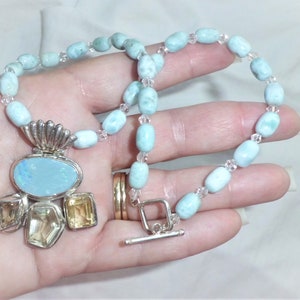 May include: A light blue gemstone necklace with a silver pendant featuring a large oval opal and smaller citrine stones. The necklace is made with oval-shaped beads and clear crystal beads.