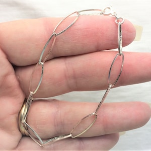 May include: A silver bracelet with an oval link design. The bracelet is made of thin, hammered metal links connected by small rings. It has a lobster clasp closure. The bracelet is shown against a neutral background.