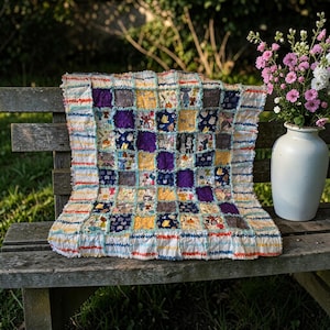 May include: A handmade patchwork quilt with a variety of patterned squares in shades of blue, yellow, purple, and gray. The quilt is placed on a wooden bench outdoors, with a vase of flowers nearby. The quilt's edges are frayed.