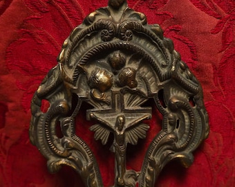 May include: Ornate bronze religious wall decor featuring a crucifix with a figure of Jesus Christ and two angels. The design is intricate and detailed, with a floral motif and a decorative border.