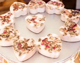 May include: Heart-shaped soaps arranged on a silver tray. The soaps are white with a floral design on top, featuring dried rose petals and herbs. The soaps are a light pink color and are arranged in a circle.
