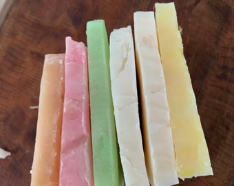 May include: An assortment of colorful cheese slices arranged on a wooden surface. The cheese varieties include shades of orange, pink, green, white, and yellow. The cheese slices are cut into rectangular shapes, showcasing a variety of textures and colors.