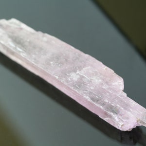 May include: A pale pink, translucent, elongated crystal with a striated surface. The crystal is approximately 4 inches long and has a slightly pointed end.