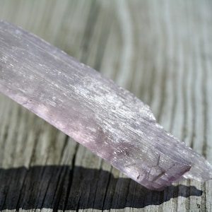 May include: A long, thin, pale purple selenite crystal with a striated surface. The crystal is lying on a wooden surface.