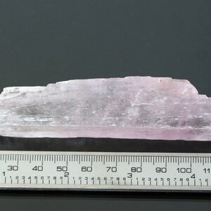 May include: A pale pink, translucent, elongated crystal with a striated surface. The crystal is approximately 6 inches long and is measured against a ruler.