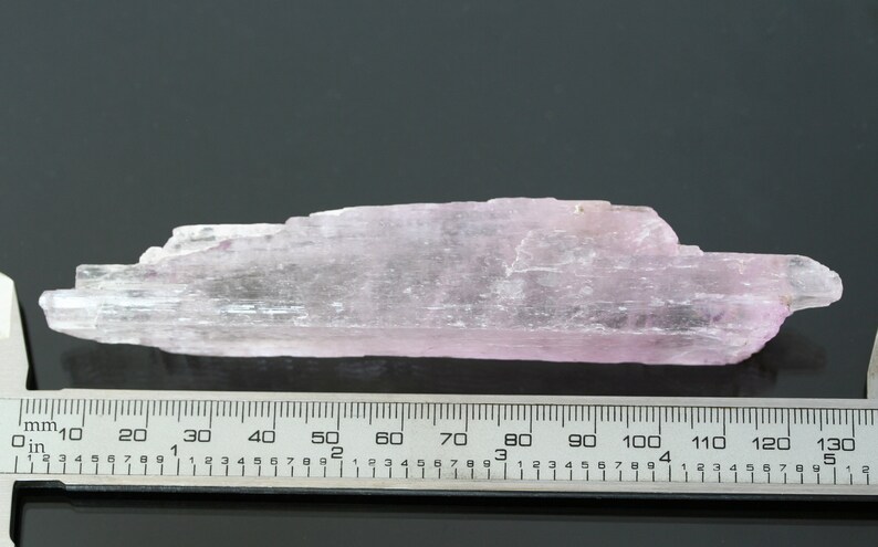 May include: A pale pink, translucent, elongated crystal with a striated surface. The crystal is approximately 6 inches long and is measured against a ruler.