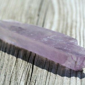 May include: A long, thin, pale purple crystal with a rough, natural texture. The crystal is lying on a light brown wooden surface.