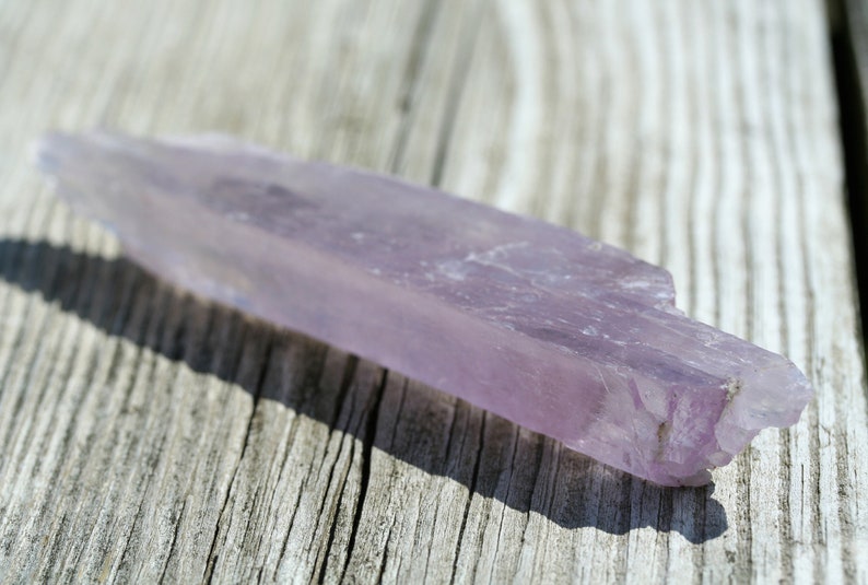 May include: A long, thin, pale purple crystal with a rough, natural texture. The crystal is lying on a light brown wooden surface.
