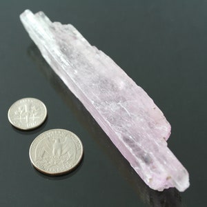 May include: A pale pink, translucent crystal with a striated texture. The crystal is about 4 inches long and is next to two US quarters for scale.