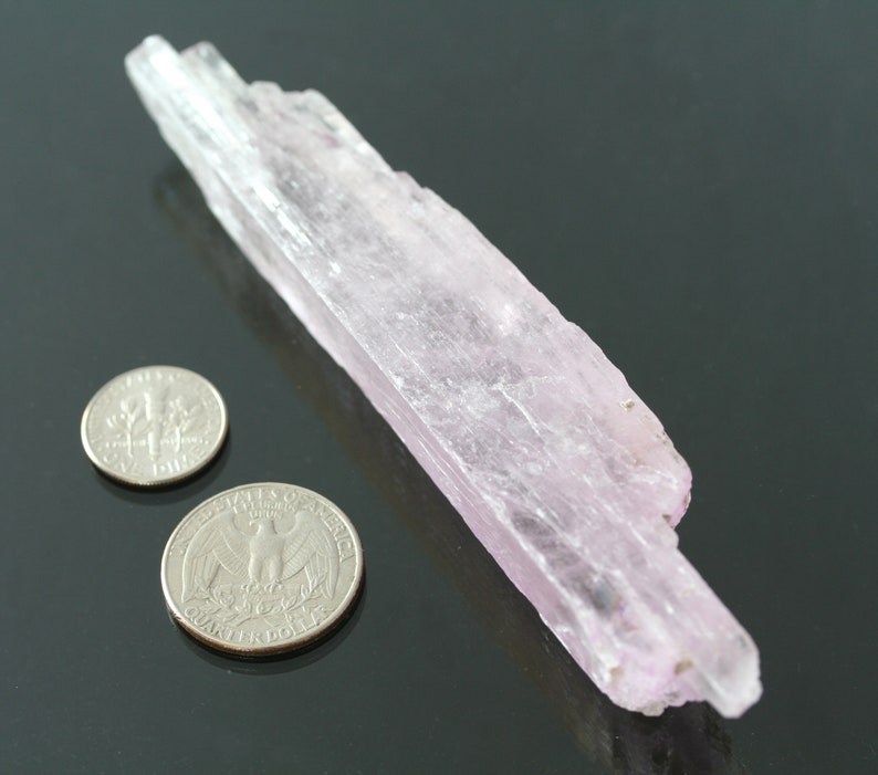 May include: A pale pink, translucent crystal with a striated texture. The crystal is about 4 inches long and is next to two US quarters for scale.