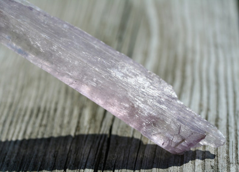 May include: A long, thin, pale purple selenite crystal with a striated surface. The crystal is lying on a wooden surface.