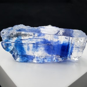 May include: A clear, crystalline mineral with vibrant blue inclusions. The rough-cut stone has a layered appearance, with streaks of deep blue running through the transparent crystal. The mineral is displayed on a white surface against a black background.