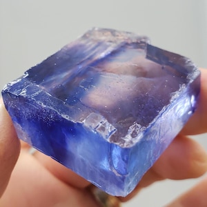 May include: A raw, natural fluorite crystal with a striking blue and purple color gradient. The crystal has a cubic shape with visible facets and a translucent quality, showcasing its internal structure. The stone is held in a hand.