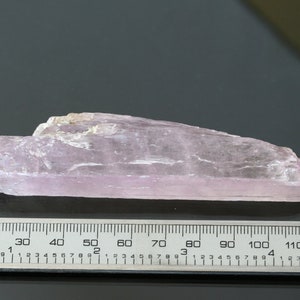 May include: A pale pink, translucent, elongated crystal with a rough, natural surface. The crystal is approximately 12 cm long and is measured against a ruler.