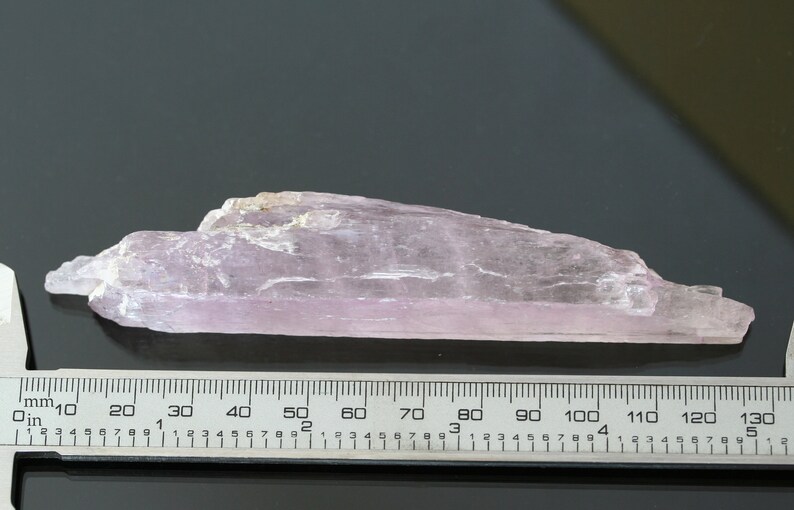 May include: A pale pink, translucent, elongated crystal with a rough, natural surface. The crystal is approximately 12 cm long and is measured against a ruler.