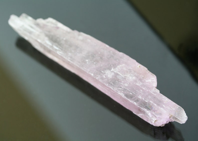 May include: A pale pink, translucent, elongated crystal with a striated surface. The crystal is approximately 4 inches long and has a slightly pointed end.