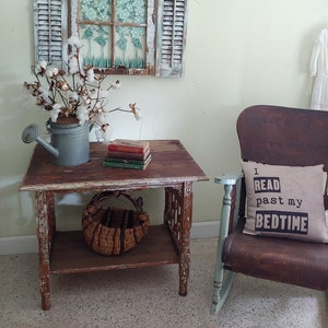 May include: A rustic wooden end table with a distressed white finish, featuring a woven basket on the lower shelf and a stack of books on the top shelf. A vintage watering can with cotton flowers sits on the table. The table is next to a rocking chair with a pillow that reads "I read past my bedtime".