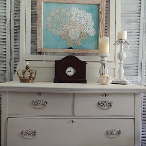 May include: White painted dresser with three drawers and a bottom shelf. The dresser has ornate metal drawer pulls. The dresser is decorated with a clock, candles, and a framed picture with doilies.