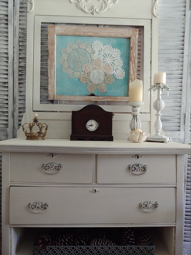 May include: White painted dresser with three drawers and a bottom shelf. The dresser has ornate metal drawer pulls. The dresser is decorated with a clock, candles, and a framed picture with doilies.