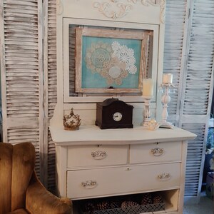 May include: White antique dresser with three drawers and a lower shelf. The dresser has ornate gold-colored hardware and sits in front of a white shuttered room divider. A small clock and a gold crown are on top of the dresser.