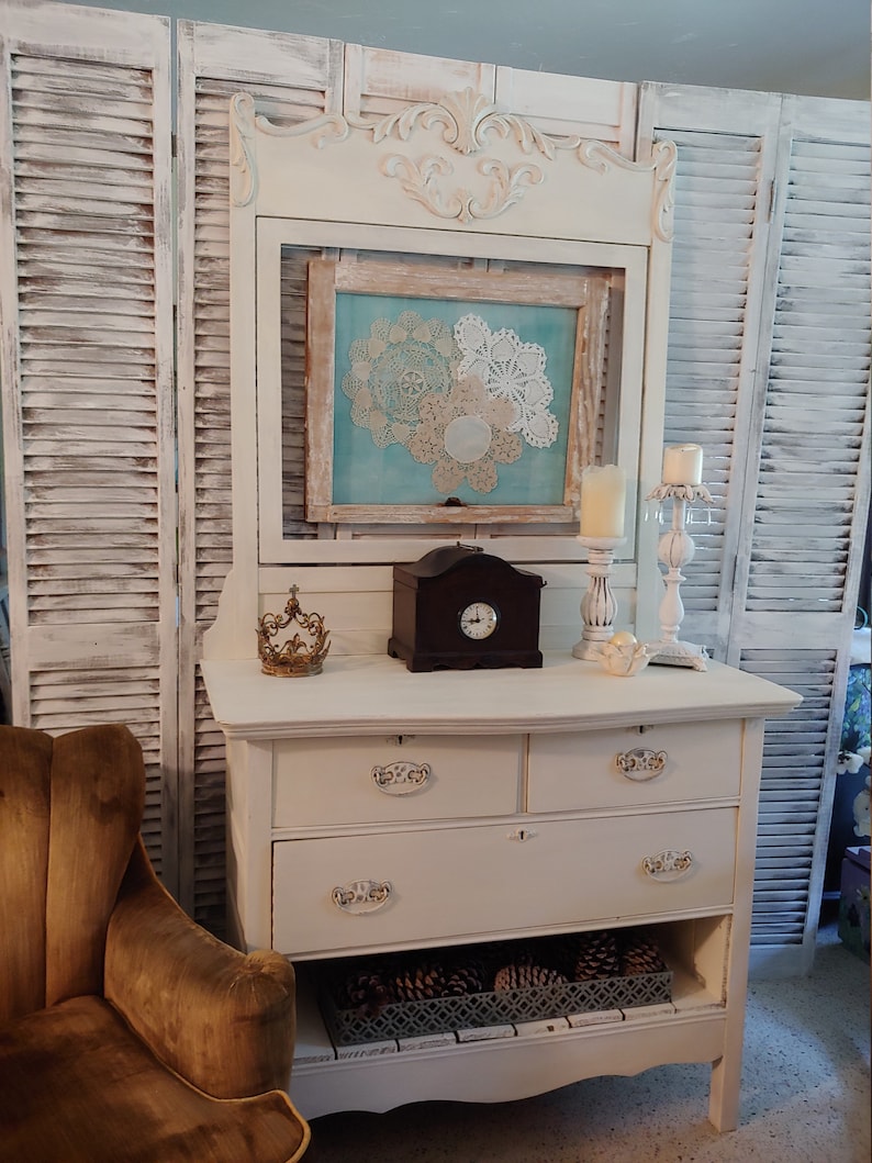 May include: White antique dresser with three drawers and a lower shelf. The dresser has ornate gold-colored hardware and sits in front of a white shuttered room divider. A small clock and a gold crown are on top of the dresser.