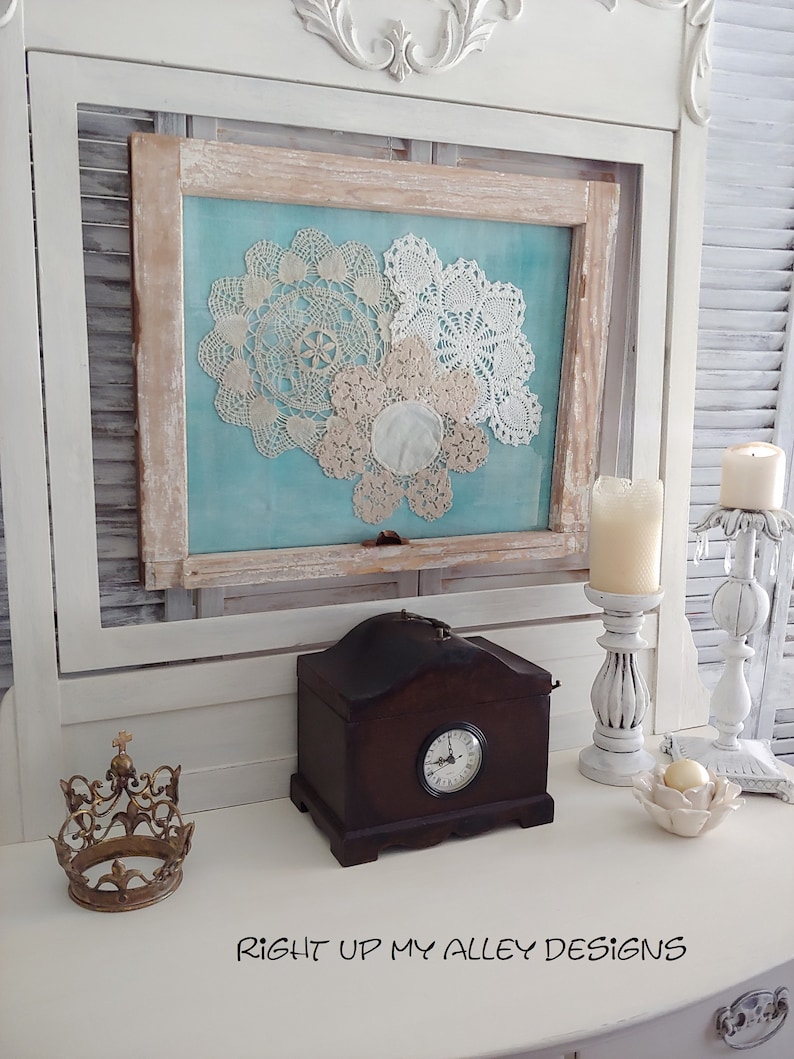 May include: A white painted wooden cabinet with a framed picture of three white doilies on a blue background. The cabinet has a small wooden clock on top and a gold crown on the bottom. The text "RIGHT UP MY ALLEY DESIGNS" is written on the surface of the cabinet.