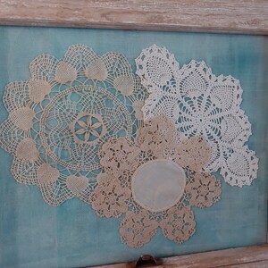May include: Three white and cream crocheted doilies displayed in a wooden frame against a teal blue background.