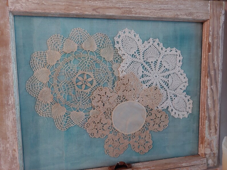 May include: Three white and cream crocheted doilies displayed in a wooden frame against a teal blue background.