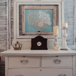 May include: White painted dresser with four drawers and ornate hardware. The dresser has a decorative top with a framed picture of doilies and a clock on top.