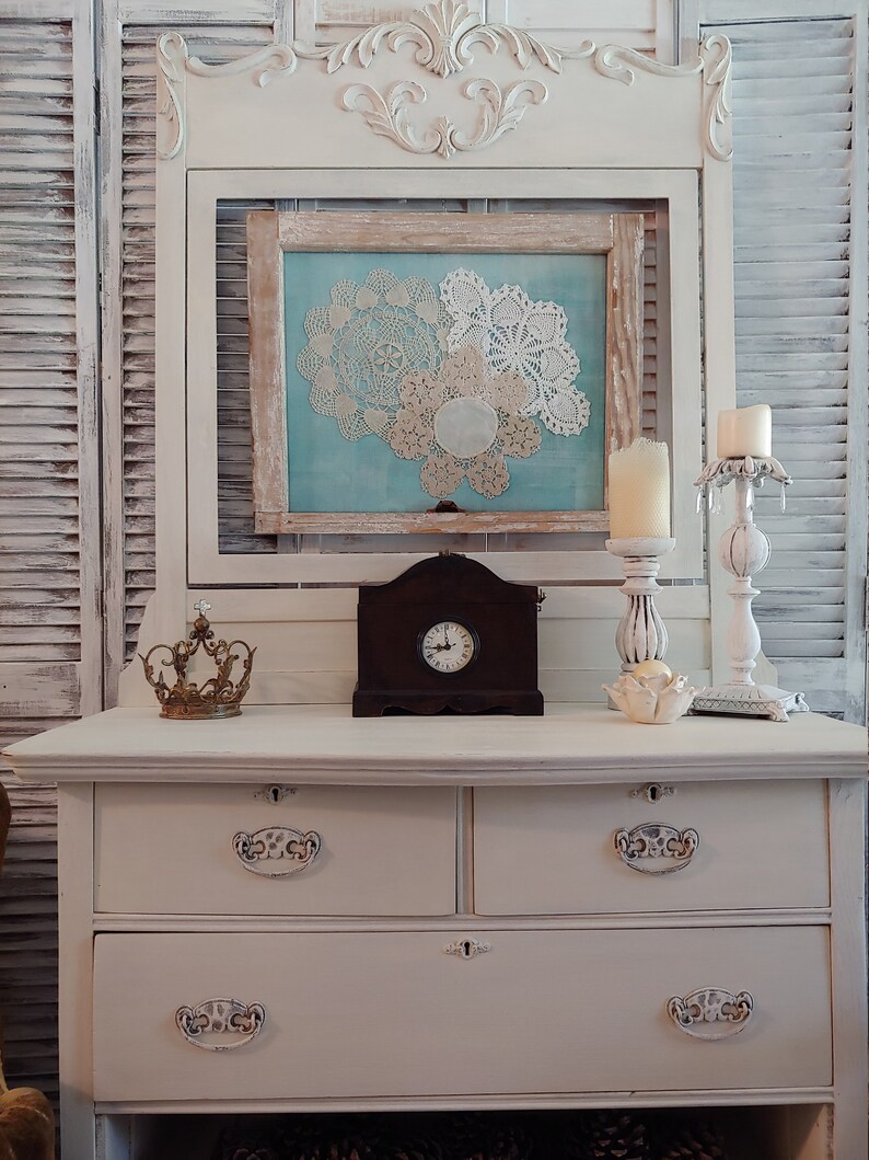 May include: White painted dresser with four drawers and ornate hardware. The dresser has a decorative top with a framed picture of doilies and a clock on top.