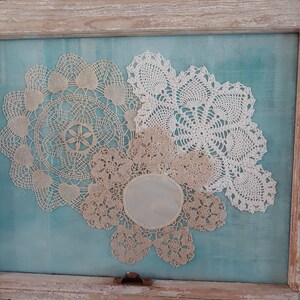 May include: Three white crocheted doilies are arranged on a turquoise blue background in a wooden frame. The doilies are different sizes and have intricate patterns.