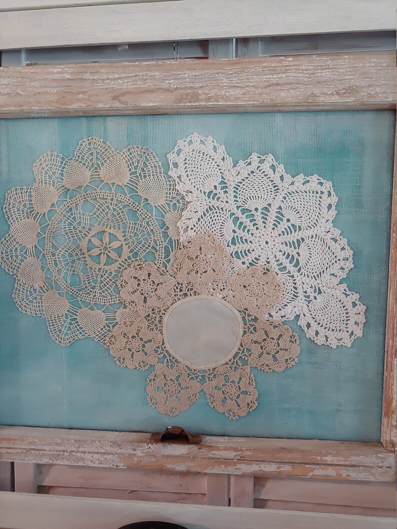 May include: Three white crocheted doilies are arranged on a turquoise blue background in a wooden frame. The doilies are different sizes and have intricate patterns.