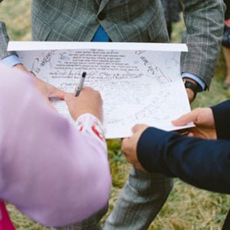 May include: A white sheet of paper with black calligraphy writing on it. Two people are signing the paper with black pens. The paper is being held by two people.