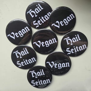 May include: A group of black circular badges with white lettering. Some badges display the word "Vegan", while others read "Hail Seitan" in a decorative font. The badges are laid out on a pale surface.