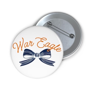 May include: Two white round buttons with a pin back. One button has the words "War Eagle" in orange script above a navy blue bow. The other button is silver.