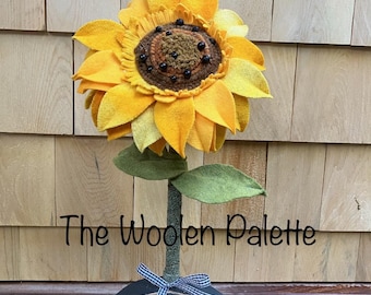 May include: A handcrafted felt sunflower with yellow petals and a brown center featuring small black beads. The flower sits on a green stem attached to a black semi-circular base with a blue and white gingham bow and a Sunflower Seeds label.