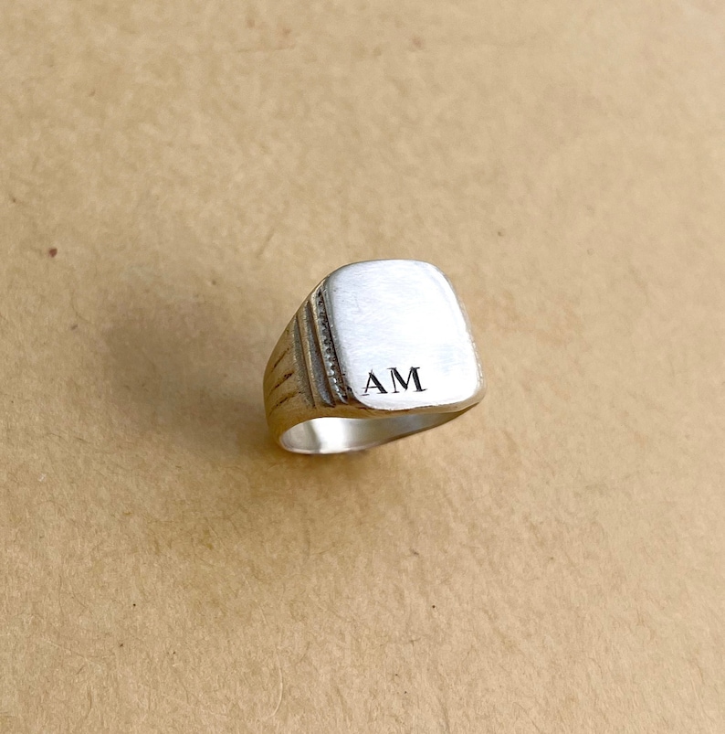 May include: A silver signet ring with a square top and the initials "AM" engraved on the top.