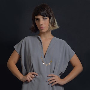 May include: A woman wearing a gray v-neck top with a gold chain necklace with multiple charms. The necklace has a key, a circle, a heart, and a leaf charm.