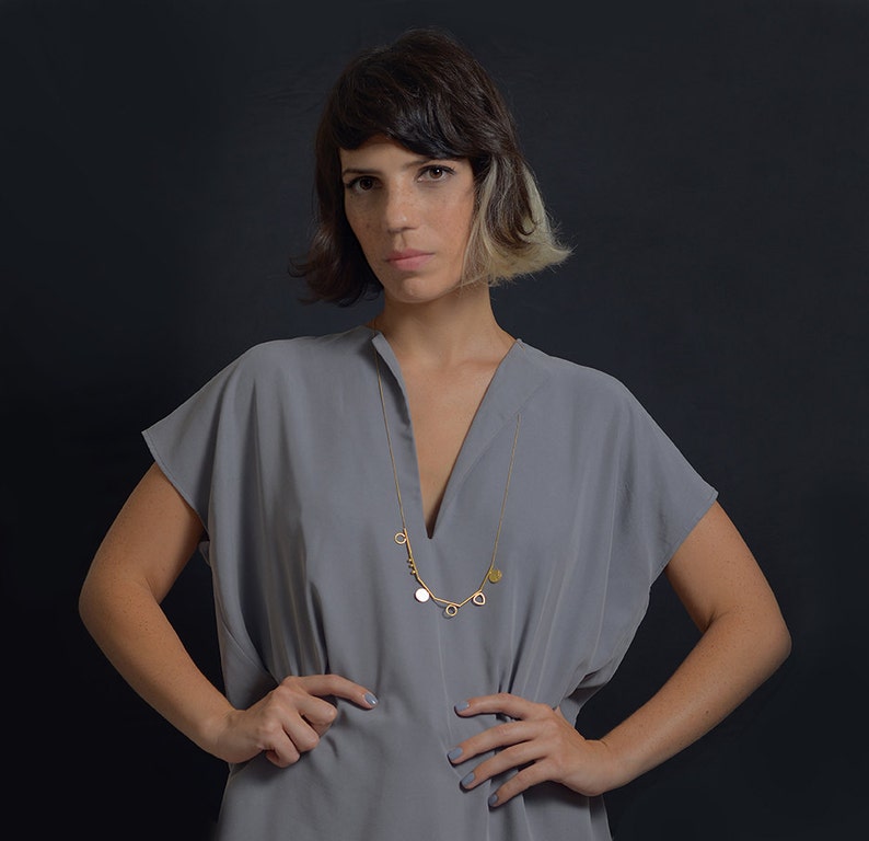 May include: A woman wearing a gray v-neck top with a gold chain necklace with multiple charms. The necklace has a key, a circle, a heart, and a leaf charm.