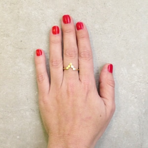 May include: A gold ring with a V-shaped design made of small circles. The ring is worn on the middle finger of a hand with red nail polish.