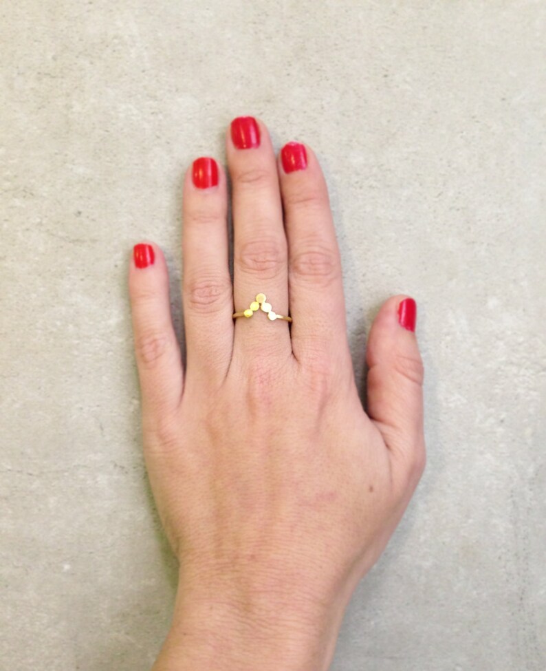May include: A gold ring with a V-shaped design made of small circles. The ring is worn on the middle finger of a hand with red nail polish.