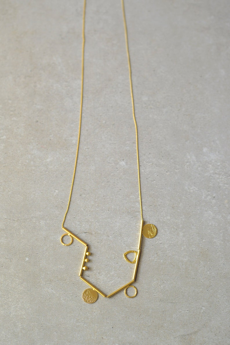 May include: A gold necklace with a geometric design featuring multiple circles, a triangle, and a bar. The necklace is made of a thin chain and has a delicate, minimalist style.