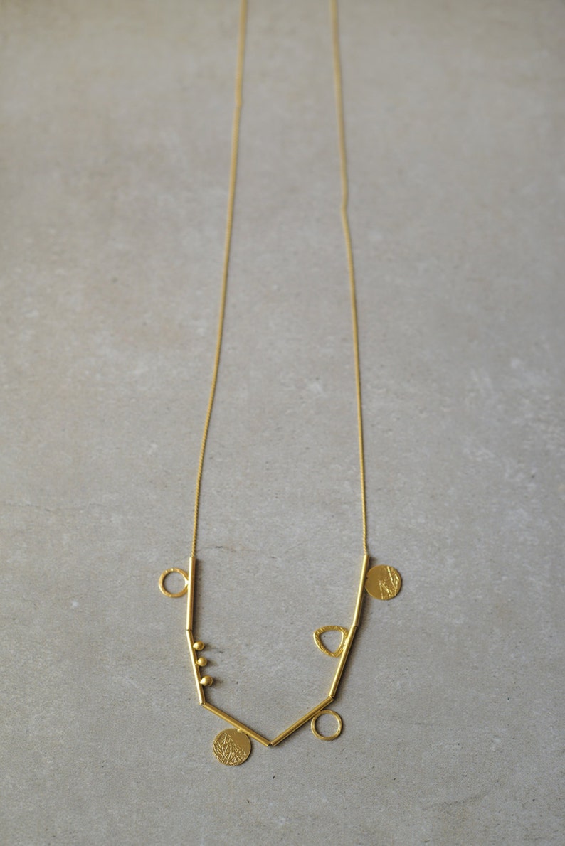 May include: A gold necklace with a geometric design featuring multiple circles, a triangle, and small beads. The necklace is made of thin gold bars connected by a delicate chain.