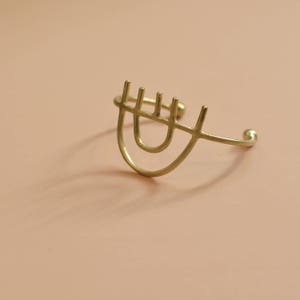 以下が含まれることがあります： 目のように見える幾何学模様が施された、金色調の金属製のブレスレット。ブレスレットは開いており、着用する準備ができています。