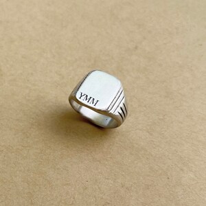 May include: A silver signet ring with a square top and a grooved band. The top of the ring is engraved with the letters "YMMI".